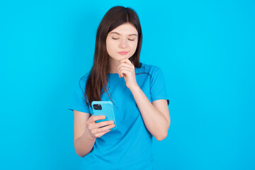 Thoughtful happy young beautiful Caucasian woman wearing blue T-shirt over blue wall hold look phone, copyspace