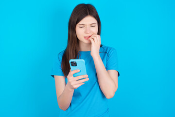 Portrait of pretty frightened young beautiful Caucasian woman wearing blue T-shirt over blue wall chatting biting nails after reading some scary news on her smartphone.