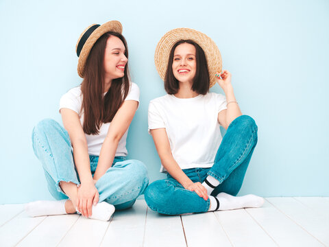 Two young beautiful smiling hipster female in trendy summer white t-shirt and jeans clothes.Sexy carefree women posing near light blue wall in studio.Positive models in hats