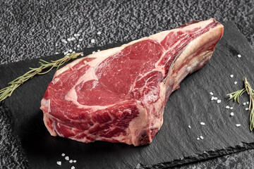 a piece of raw beef steak with a bone lies on a black stone background with a decor of dry greens