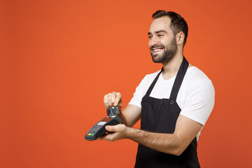 Young man barista bartender barman employee in apron white t-shirt work in coffee shop use wireless bank payment terminal acquire credit card payment isolated on orange background. Business startup.
