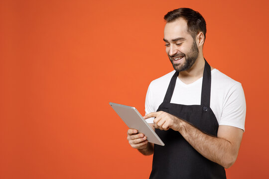 Young smiling fun man 20s barista bartender barman employee in black apron white t-shirt work in coffee shop using tablet pc computer browsing internet isolated on orange background. business startup.