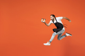 Fototapeta na wymiar Full length side view young man barista bartender barman employee in apron white tshirt work in coffee shop jump hold paper delivery cup run fast isolated on orange background. Small business startup