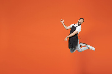 Full length young fun man barista bartender barman employee in black apron white tshirt work in coffee shop jump high play guitar gesture isolated on orange background. Small business startup concept.