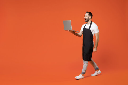 Full Length Young Man Barista Bartender Barman Employee In Black Apron White T-shirt Work In Coffee Shop Using Laptop Pc Computer Walk Isolated On Orange Background. Small Business Startup Concept.