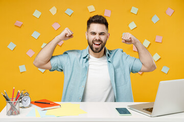 Young strong employee business man 20s in shirt sit work at white office desk with pc laptop showing biceps muscles on hand demonstrating strength power isolated on yellow background studio portrait