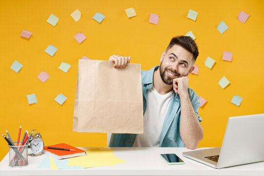 Young dreamful employee business man in shirt sit work at white office desk pc laptop hold brown clear blank craft paper takeaway bag mock up slook aside prop up chin isolated on yellow background