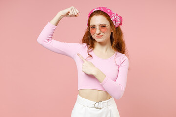 Obraz na płótnie Canvas Young strong sporty fitness woman in rose clothes bandana glasses point index finger on biceps muscles on hand demonstrating strength power isolated on pastel pink background studio Lifestyle concept