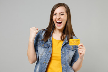 Young fun happy caucasian woman 20s in casual trendy denim jacket yellow t-shirt holding in hand credit bank card look camera clench fist do winner gesture isolated on grey background studio portrait