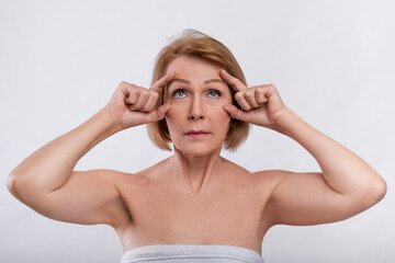 Portrait of senior lady dissatisfied with wrinkles around her eyes, touching her face on light studio background