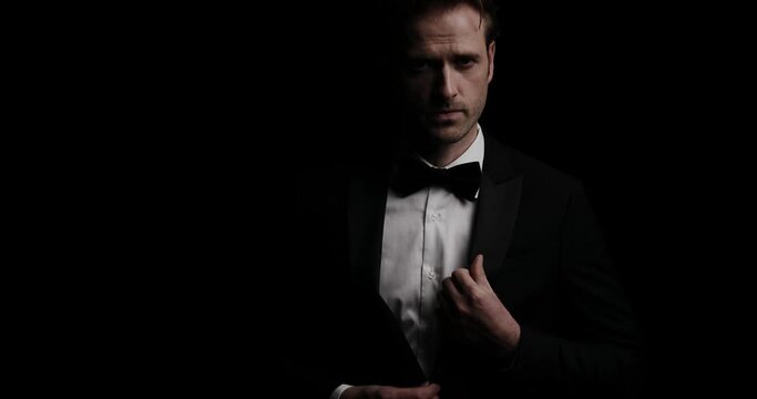 handsome businessman with attitude is arranging his tux, looking down, rubbing his palms and smiling on dark background