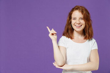 Young friendly happy smiling caucasian woman 20s wearing white basic t-shirt pointing index finger aside on workspace area mock up copy space isolated on dark violet color background studio portrait