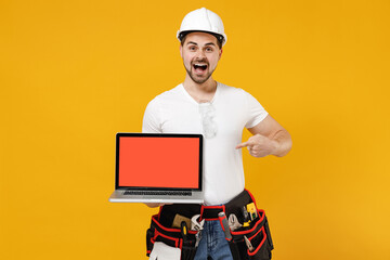 Young employee handyman man in protective helmet hardhat point finger on laptop pc computer blank screen workspace area isolated on yellow background. Instruments apartment room. Repair home concept.