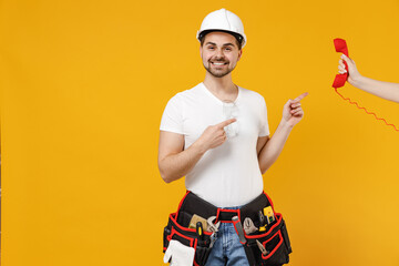 Young employee handyman man in protective helmet hardhat point finger on hand with handset phone isolated on yellow background Instruments accessories renovation apartment room Repair home concept