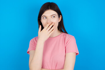 Emotional young beautiful Caucasian woman wearing pink T-shirt over blue wall gasps from astonishment, covers opened mouth with palm, looks shocked at camera. Reaction concept