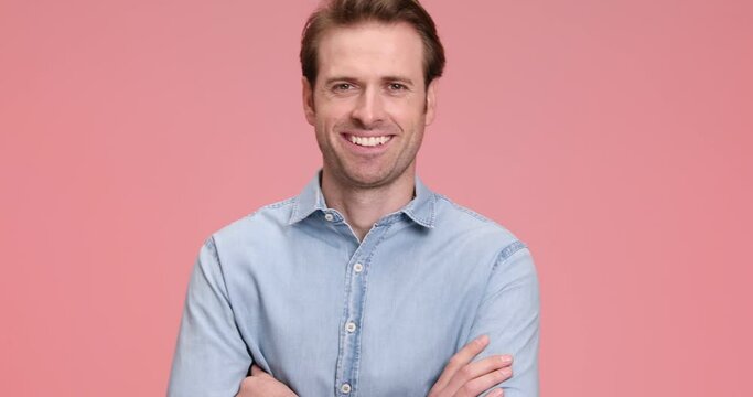 smiling attractive man in blue jeans shirt folding arms while camera was approaching, laughing and posing on pink background in studio