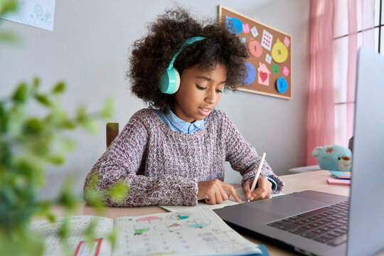 Focused preteen black African American girl wear headphones with laptop sitting writing listening tutor online virtual lesson studying at home doing task. Remote distant homeschooling concept.