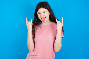 young beautiful Caucasian woman wearing pink T-shirt over blue wall making rock hand gesture and...