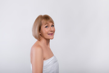 Anti-aging care. Attractive senior woman in towel pampering her sensitive mature skin on light studio background