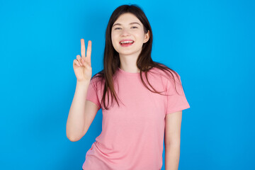 young beautiful Caucasian woman wearing pink T-shirt over blue wall showing and pointing up with fingers number two while smiling confident and happy.
