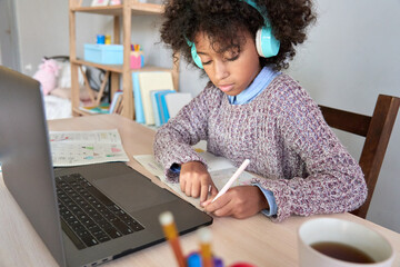 Concentrated preteen black African American girl wearing headphones sitting writing listening to...