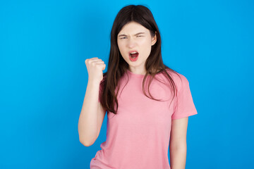young beautiful Caucasian woman wearing pink T-shirt over blue wall angry and mad raising fist frustrated and furious while shouting with anger. Rage and aggressive concept.