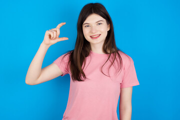 young beautiful Caucasian woman wearing pink T-shirt over blue wall smiling and confident gesturing with hand doing small size sign with fingers looking and the camera. Measure concept