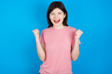 young beautiful Caucasian woman wearing pink T-shirt over blue wall celebrating surprised and amazed for success with arms raised and open eyes. Winner concept.