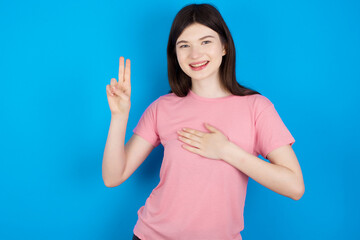 young beautiful Caucasian woman wearing pink T-shirt over blue wall smiling swearing with hand on...