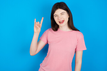 Fototapeta premium young beautiful Caucasian woman wearing pink T-shirt over blue wall doing a rock gesture and smiling to the camera. Ready to go to her favorite band concert.