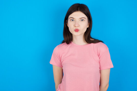 Shot of pleasant looking young beautiful Caucasian woman wearing pink T-shirt over blue wall, pouts lips, looks at camera, Human facial expressions