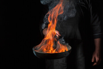  Chef cooking food in pan with fire flame on black background. Restaurant and hotel service...