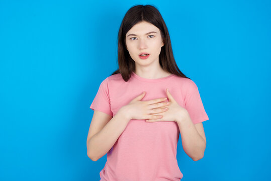 Scared young beautiful Caucasian woman wearing pink T-shirt looks with frightened expression, keeps hands on chest, being puzzled to notice something strange, People, hush reaction and emotions.