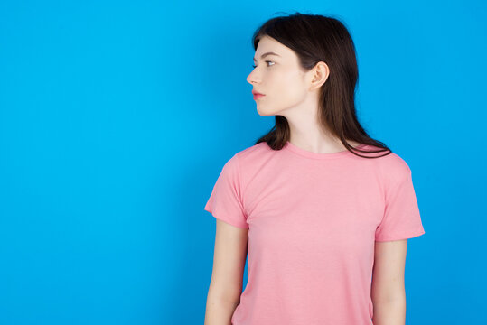 Close up side profile photo young beautiful Caucasian woman wearing pink T-shirt over blue wall not smiling attentive listen concentrated