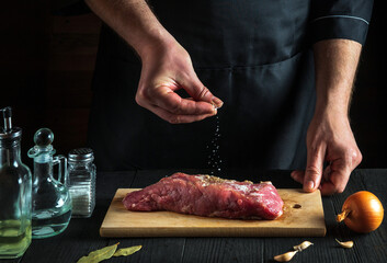 Chef or cook sprinkles raw meat with salt. Preparing beef meat before baking. Working environment in the kitchen of restaurant or cafe