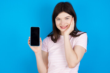 Fototapeta premium young beautiful Caucasian woman wearing stripped T-shirt over blue wall hold hand modern technology use touch face palm astonished impressed scream wow omg unbelievable unexpected