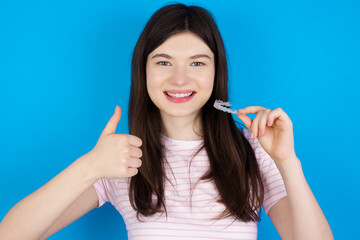young beautiful Caucasian woman wearing stripped T-shirt over blue wall holding an invisible braces aligner and rising thumb up, recommending this new treatment. Dental healthcare concept.