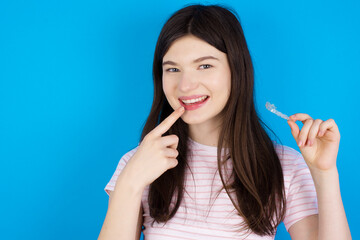 young beautiful Caucasian woman wearing stripped T-shirt over blue wall holding an invisible aligner and pointing to her perfect straight teeth. Dental healthcare and confidence concept.