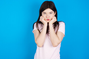 young beautiful Caucasian woman wearing stripped T-shirt over blue wall with surprised expression keeps hands under chin keeps lips folded makes funny grimace