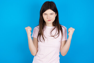 Fototapeta na wymiar Irritated young beautiful Caucasian woman wearing stripped T-shirt over blue wall blows cheeks with anger and raises clenched fists expresses rage and aggressive emotions. Furious model