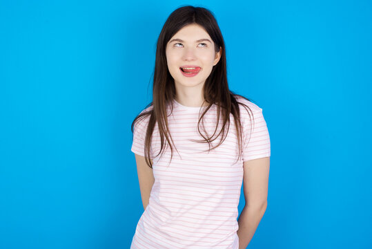 Funny young beautiful Caucasian woman wearing stripped T-shirt over blue wall makes grimace and crosses eyes plays fool has fun alone sticks out tongue.