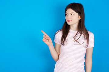 young beautiful Caucasian woman wearing stripped T-shirt over blue wall points at copy space and advertises something, advices best price.