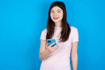 young beautiful Caucasian woman wearing stripped T-shirt over blue wall holds mobile phone in hands and rejoices positive news, uses modern cellular