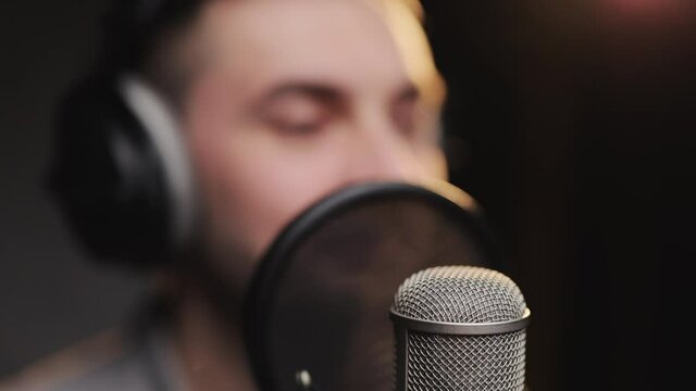Caucasian mixed man singer wearing headphones is performing a song with a microphone while recording in music studio with low lights