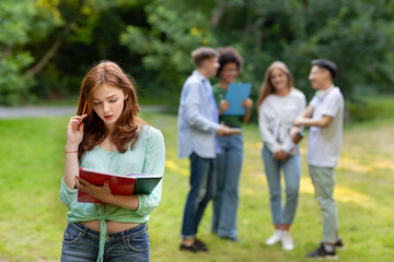 Focused redhead student girl preparing for lecture outdoors while her groupmates chatting