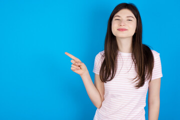 Positive young beautiful Caucasian woman wearing stripped T-shirt over blue wall with satisfied expression indicates at upper right corner shows good offer suggests to click on link