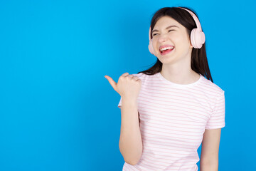 young beautiful Caucasian woman wearing stripped T-shirt over blue wall listens audio track via wireless headphones points thumb away advertises copy space