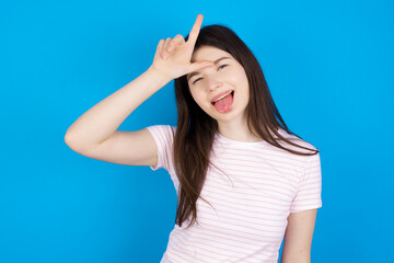 Funny young beautiful Caucasian woman wearing stripped T-shirt over blue wall makes loser gesture mocking at someone sticks out tongue making grimace face.