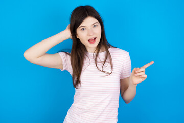 Fototapeta premium Surprised young beautiful Caucasian woman wearing stripped T-shirt over blue wall pointing at empty space holding hand on head