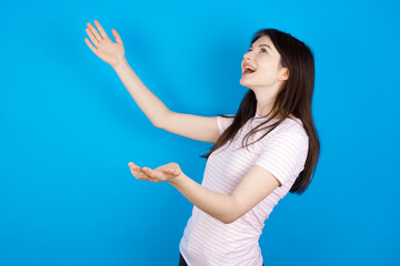 Fototapeta premium Funny astonished young beautiful Caucasian woman wearing stripped T-shirt over blue wall look empty space with arms opened ready to catch something.